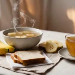 A cozy, minimalistic setting featuring stomach flu-friendly foods and drinks: a warm bowl of chicken broth, a banana, plain toast, and a cup of ginger tea, placed on a soft napkin with warm lighting to create a calm, healing atmosphere